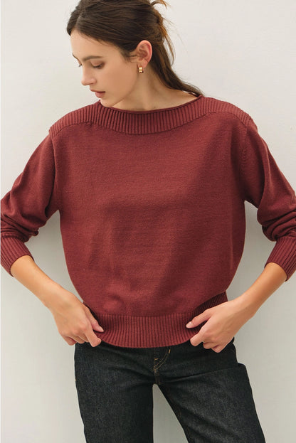 A woman wearing a rust-red knit sweater with a wide boat neckline and ribbed cuffs, paired with dark jeans, posing against a light gray wall.