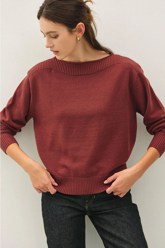 A woman wearing a rust-red knit sweater with a wide boat neckline and ribbed cuffs, paired with dark jeans, posing against a light gray wall.