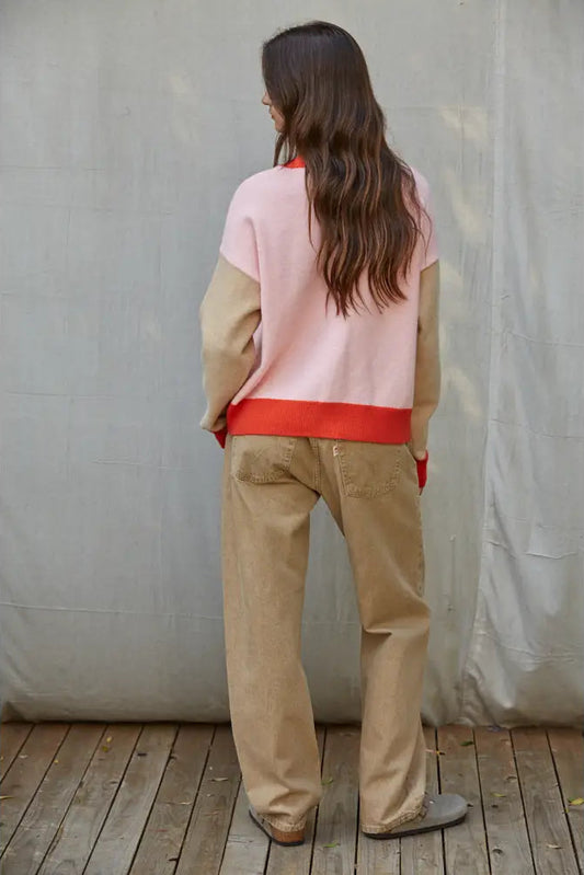 A woman viewed from behind wearing a color-blocked knit sweater with a light pink upper section, beige sleeves, and a bright red waistband, paired with beige trousers and gray slip-on shoes, standing against a gray fabric backdrop on a wooden floor.