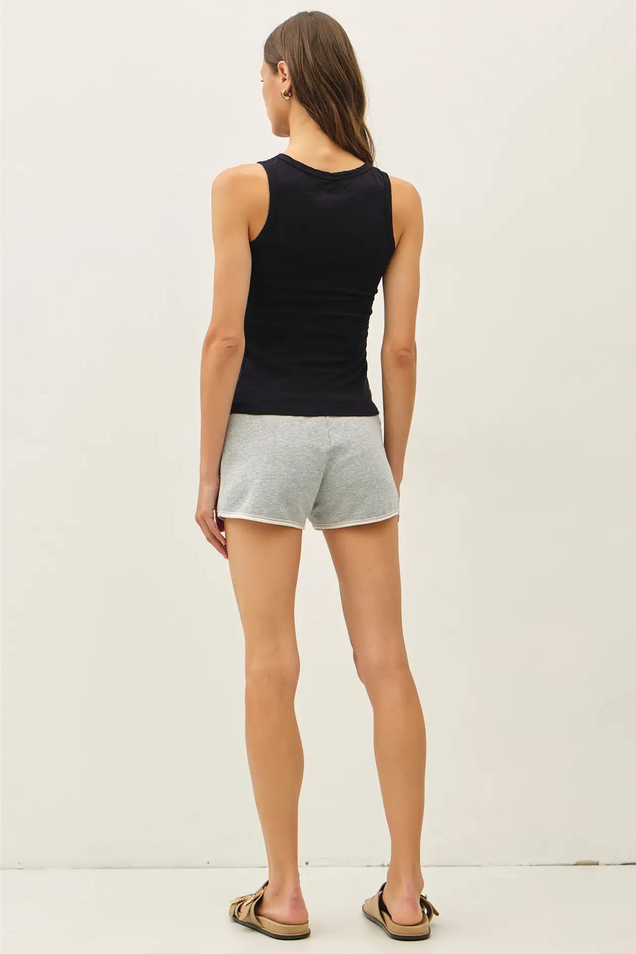 A woman shown from the back wearing a black ribbed tank top and light gray heathered shorts, standing against a plain off-white studio background.