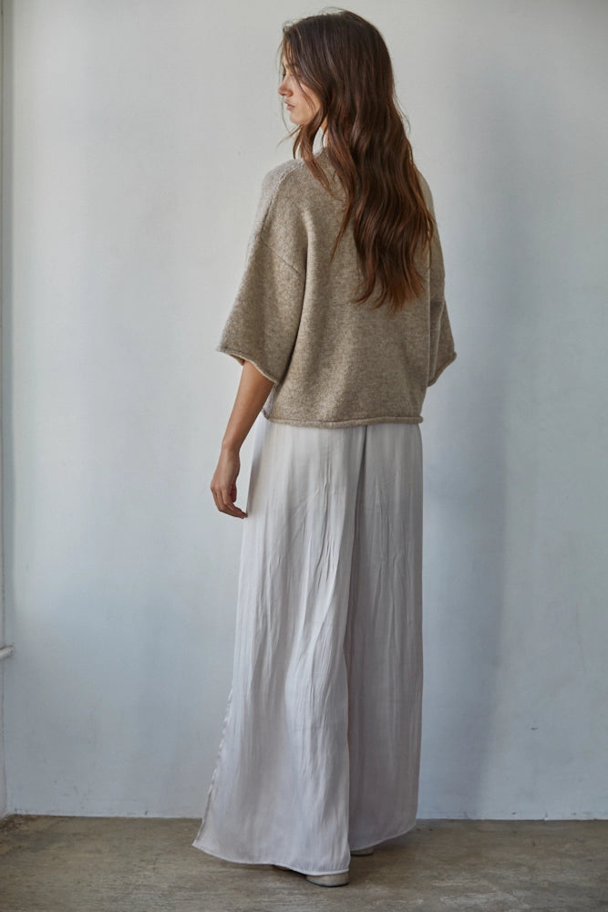 Woman wearing a beige sweater and light gray pants against a plain white wall.