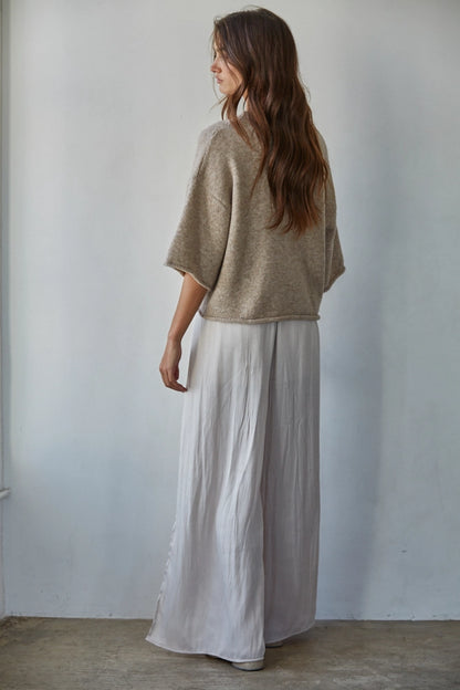 Woman wearing a beige sweater and light gray pants against a plain white wall.