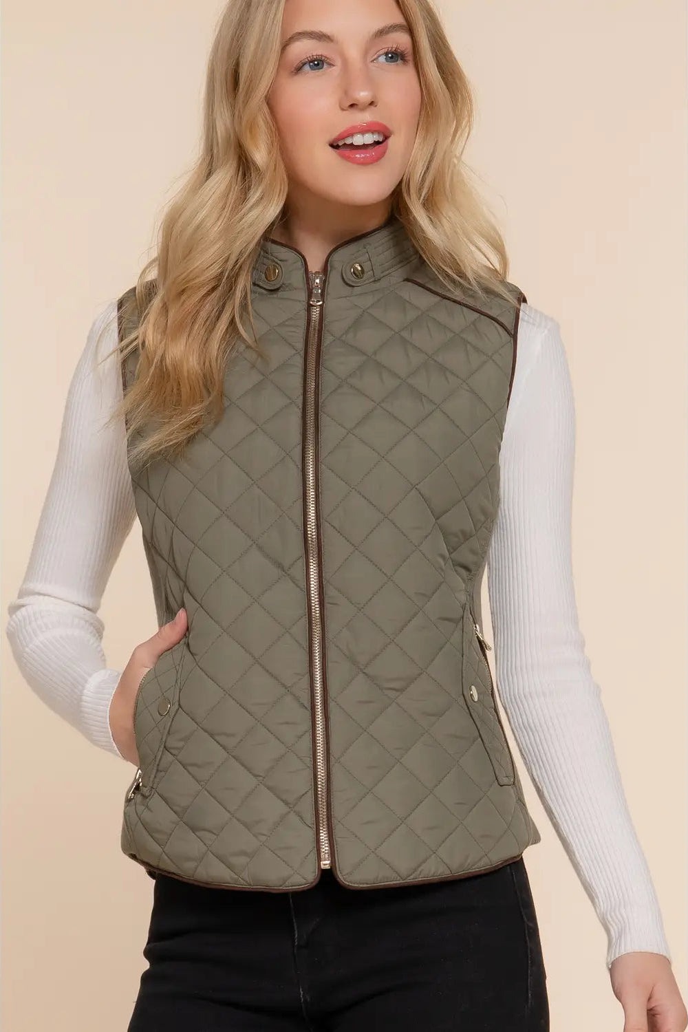A woman wearing an olive green diamond-quilted zip-front vest over a white ribbed long-sleeve top and black pants, photographed against a plain beige studio background.