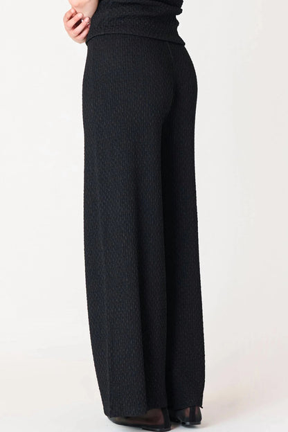 A person wearing black textured knit wide-leg pants and a matching black top, shown from the back against a plain light background.
