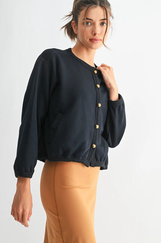 A woman wearing a dark navy blue cropped cardigan with gold buttons and tan pants, photographed against a clean white studio background.