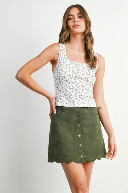 A woman modeling a sleeveless white tank top with a small multicolor floral print and an olive green mini skirt with a scalloped hem and front button closure, against a light gray studio background.