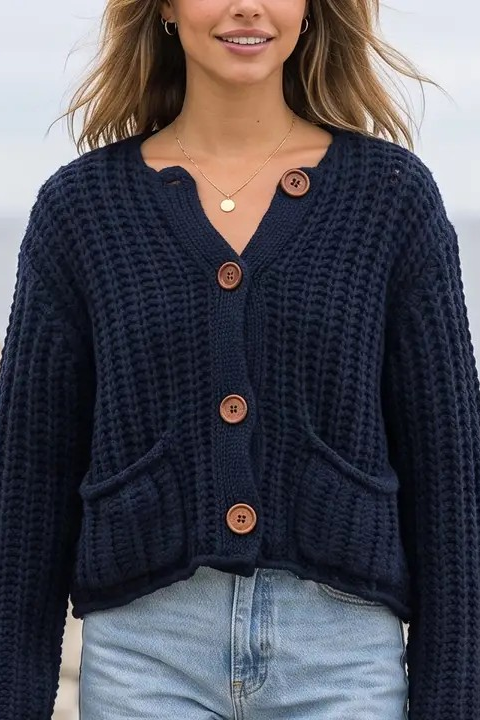 A woman wearing a navy blue waffle-knit cropped cardigan with brown wooden buttons, paired with light blue jeans, standing outdoors with a blurred rocky and ocean background.