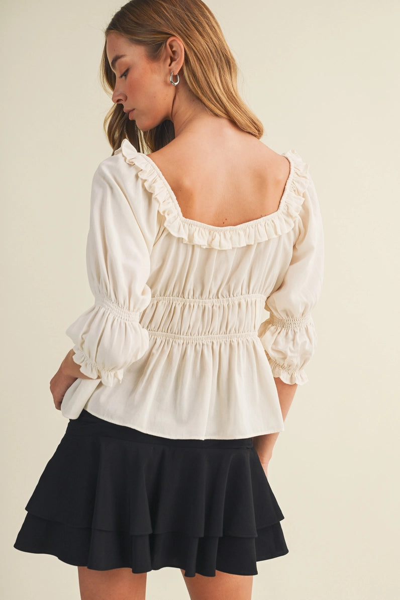 A woman wearing a cream puff-sleeve blouse with a smocked back and ruffled neckline, paired with a black ruffled mini skirt, shown from a rear three-quarter angle against a light beige background.