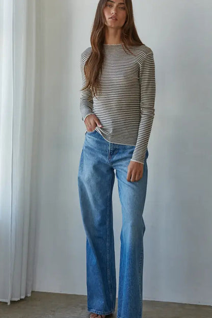 A woman modeling a beige and gray striped long-sleeve knit top with wide-leg blue jeans, standing in a bright room with white walls and a curtain.