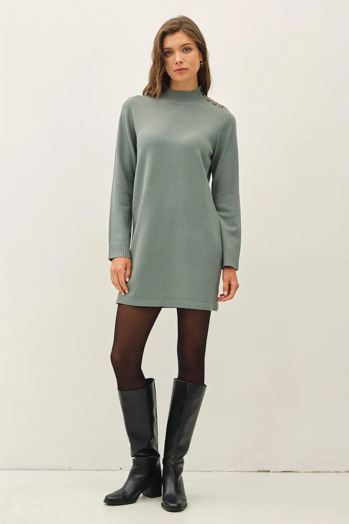 A woman modeling a sage green mock-neck knit sweater dress with decorative buttons on the shoulders, paired with black tights and black leather knee-high boots, against a plain off-white studio background.