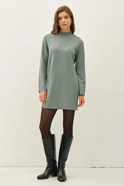 A woman modeling a sage green mock-neck knit sweater dress with decorative buttons on the shoulders, paired with black tights and black leather knee-high boots, against a plain off-white studio background.