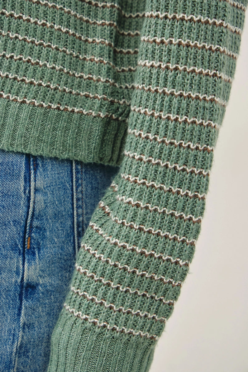 Close-up of a green ribbed knit sweater with thin beige horizontal stripes, shown paired with blue jeans against a light neutral background.