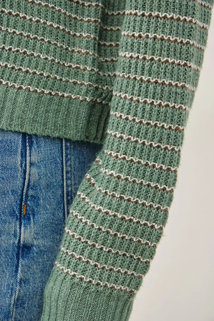 Close-up of a green ribbed knit sweater with thin beige horizontal stripes, shown paired with blue jeans against a light neutral background.