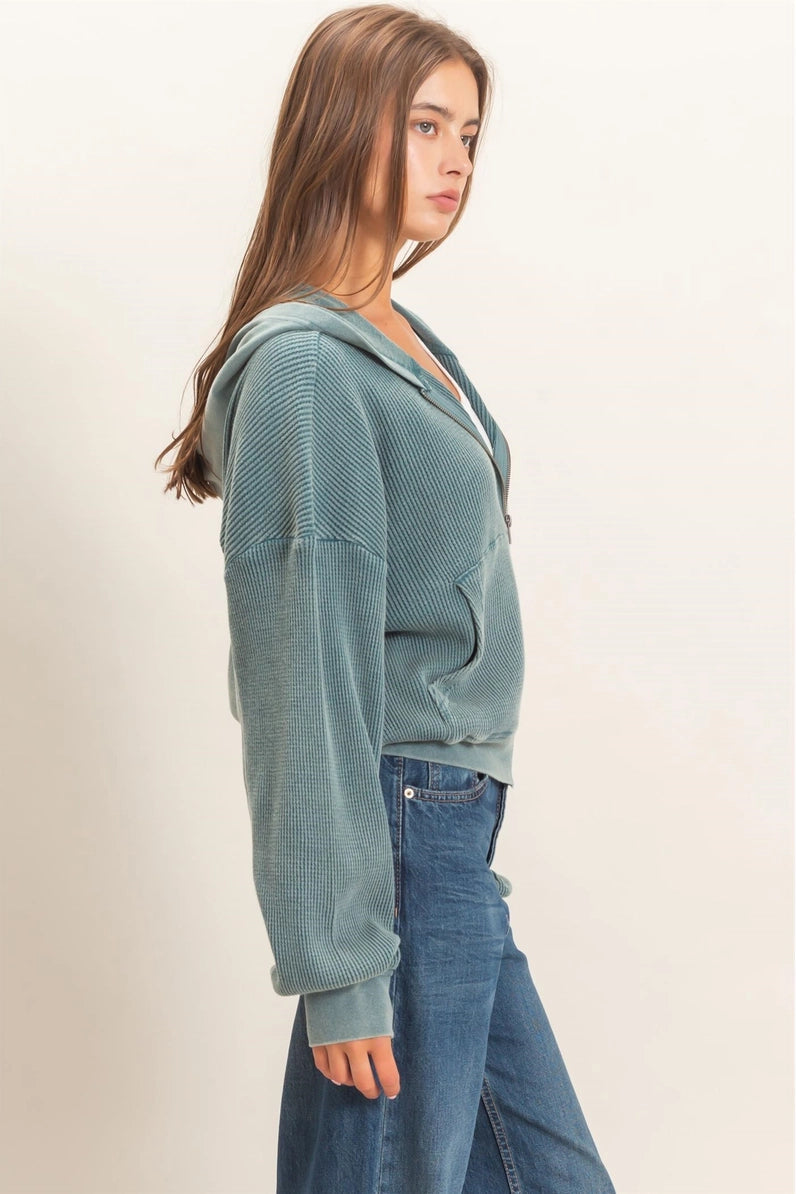 A woman shown in side profile wearing a textured, ribbed knit zip-up hoodie in a muted sage green color, paired with blue jeans, against a plain off-white studio background.