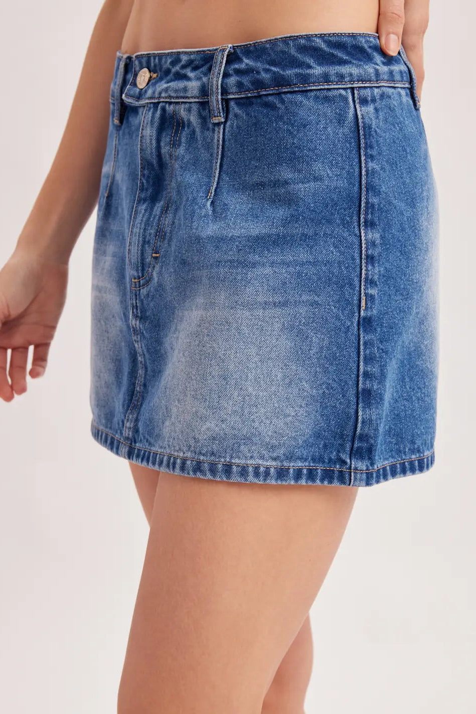 A close-up of a model wearing a medium wash blue denim mini skirt with a faded wash effect, shown from the waist down against a plain light background.