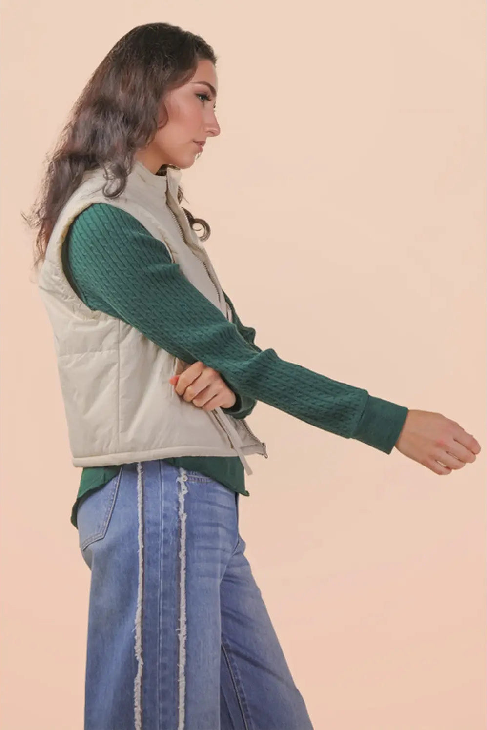 A woman shown in side profile wearing a light beige quilted puffer vest over a dark green textured long-sleeve top and blue jeans, set against a solid pale peach background.