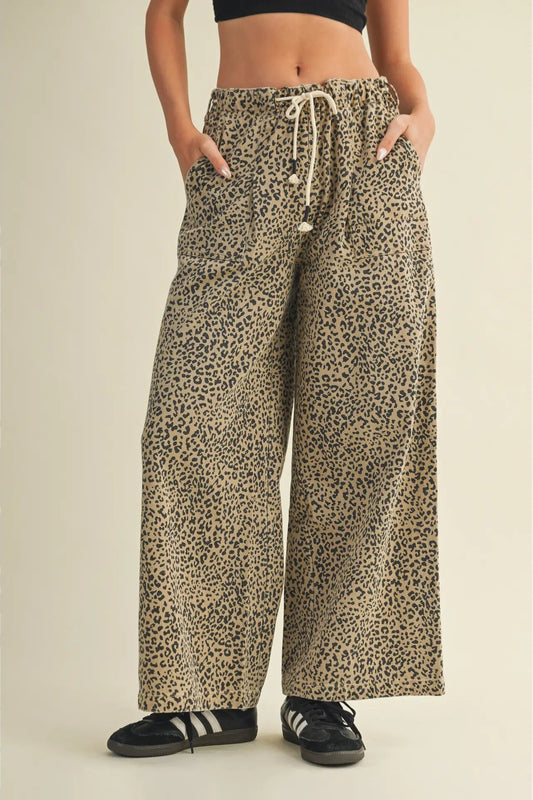 A person wearing loose, wide-leg leopard-print pants with a drawstring waist, shown from the torso down against a plain beige studio background.