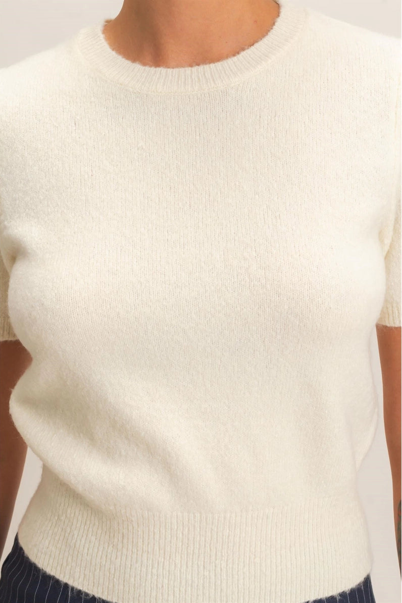 A close-up of a person wearing a short-sleeve crew-neck knit sweater in off-white or cream, paired with dark pinstriped bottoms.