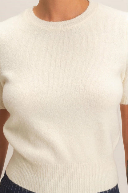 A close-up of a person wearing a short-sleeve crew-neck knit sweater in off-white or cream, paired with dark pinstriped bottoms.
