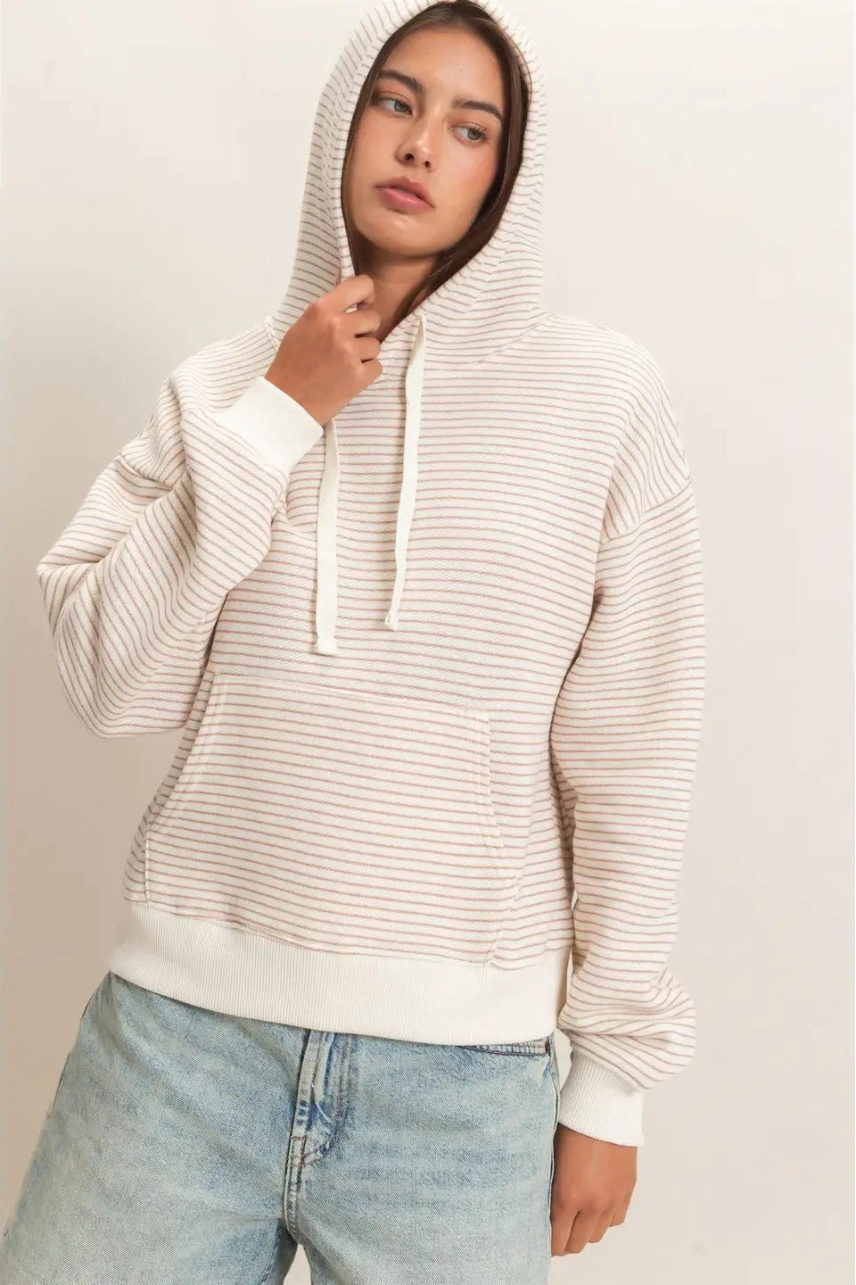 A woman wearing a cream and white horizontally striped pullover hoodie with white drawstrings and a front kangaroo pocket, paired with light-wash blue jeans, set against a plain light beige studio background.
