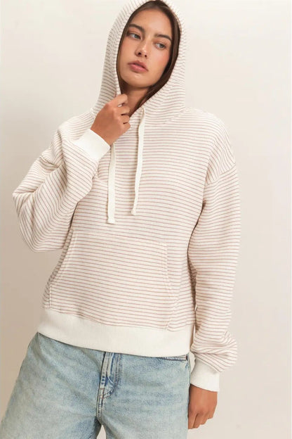 A woman wearing a cream and white horizontally striped pullover hoodie with white drawstrings and a front kangaroo pocket, paired with light-wash blue jeans, set against a plain light beige studio background.