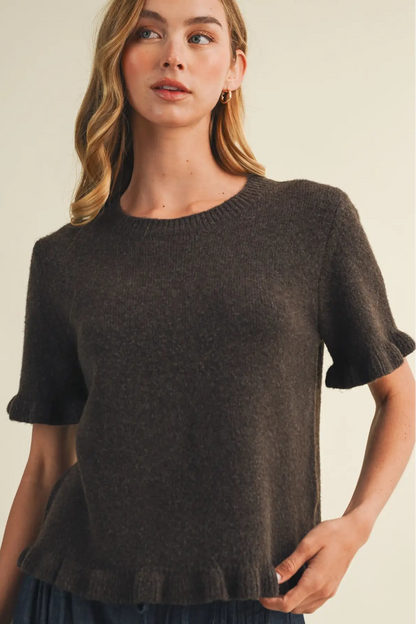 A woman wearing a dark brown short-sleeve knit sweater with a ruffled hem and sleeve cuffs, photographed against a plain light beige studio background.