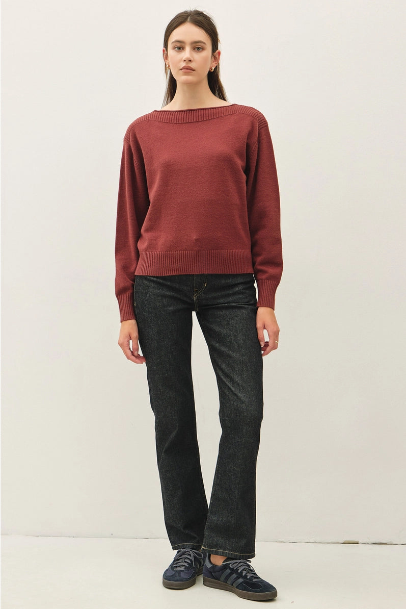 A woman modeling a rust-red knit sweater with dark charcoal jeans and navy sneakers, standing against a plain off-white studio background.