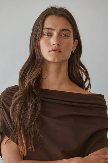 A woman with long brown hair wearing a dark brown off-the-shoulder draped top, photographed against a plain light gray background.