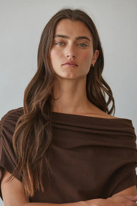 A woman with long brown hair wearing a dark brown off-the-shoulder draped top, photographed against a plain light gray background.