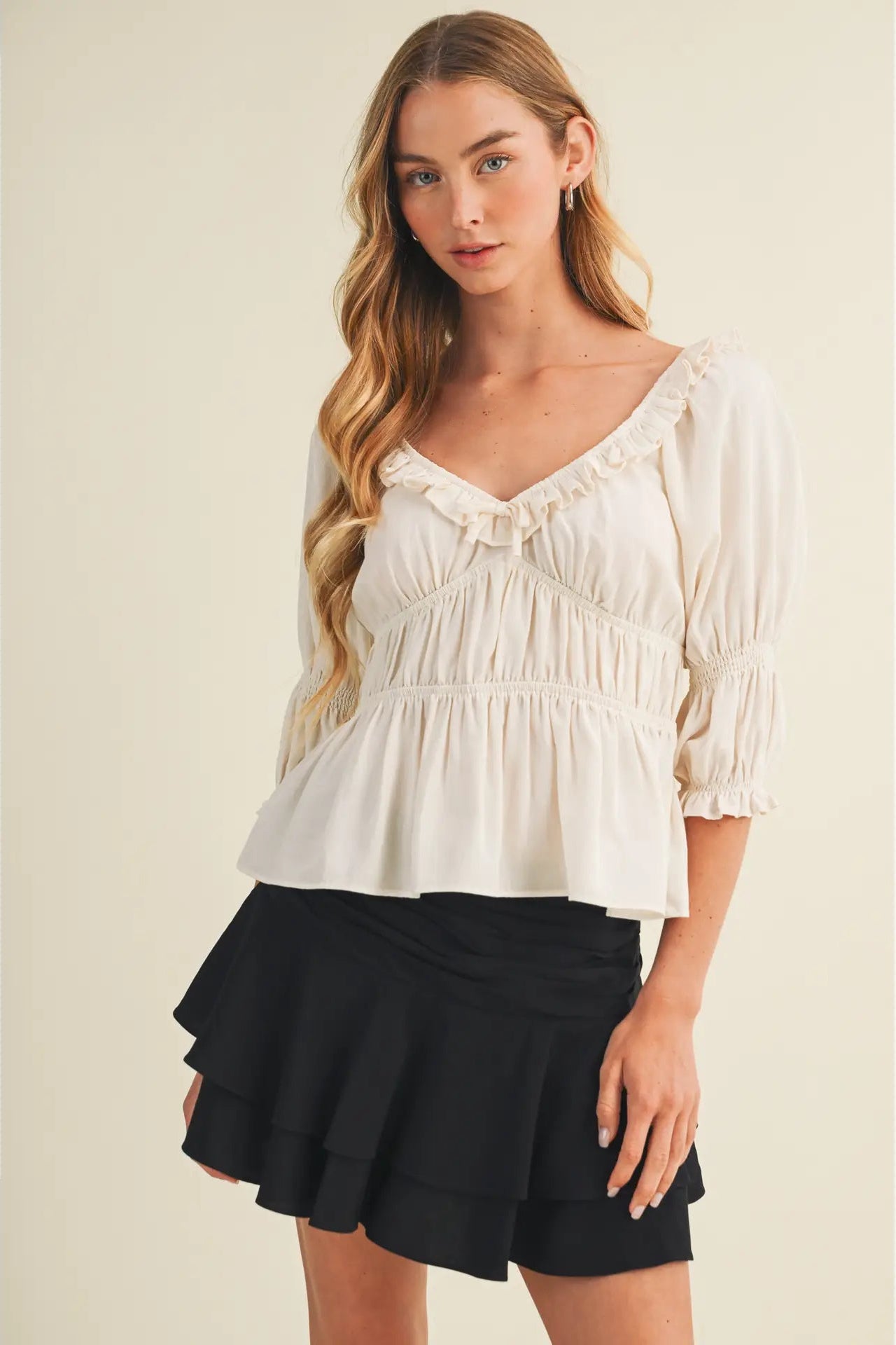 A woman modeling a cream-colored ruffled blouse with puff sleeves and a black ruffled mini skirt, set against a plain beige studio background.