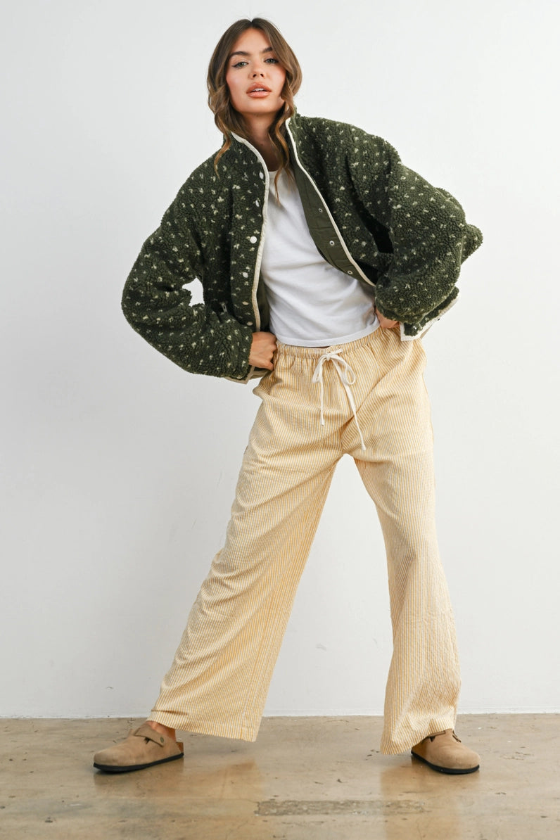 A model posing in a studio setting wearing a dark green patterned bomber jacket, a white t-shirt, cream wide-leg corduroy pants, and brown suede loafers.