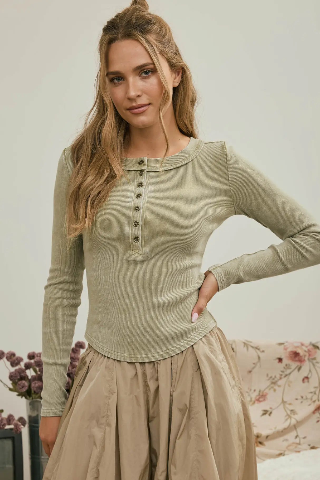 A woman modeling a light sage green long-sleeve henley top with a button placket, paired with a beige skirt, in a softly lit indoor setting.