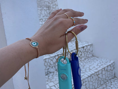 Hand holding a set of keys with a blue and gold keychain, wearing a gold bracelet with an eye design.