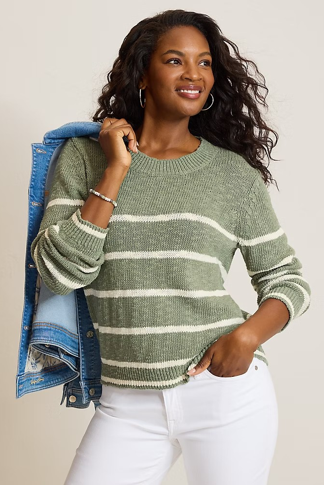 A woman wearing a sage green and white striped knit sweater, styled with a blue denim jacket draped over her shoulder and white pants, against a plain light beige background.