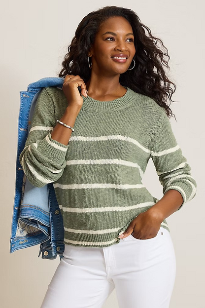 A woman wearing a sage green and white striped knit sweater, styled with a blue denim jacket draped over her shoulder and white pants, against a plain light beige background.