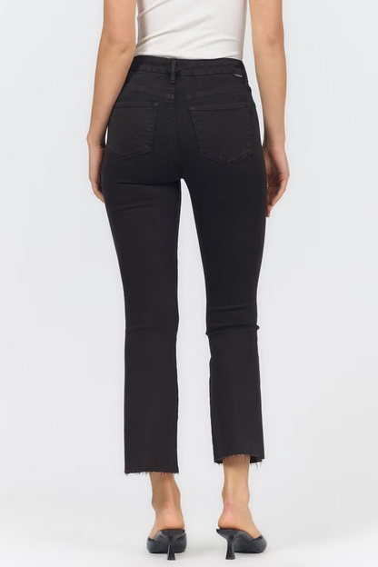 Rear view of a model wearing black cropped ankle jeans with a frayed hem, paired with a white top and black mule heels, against a light gray studio background.
