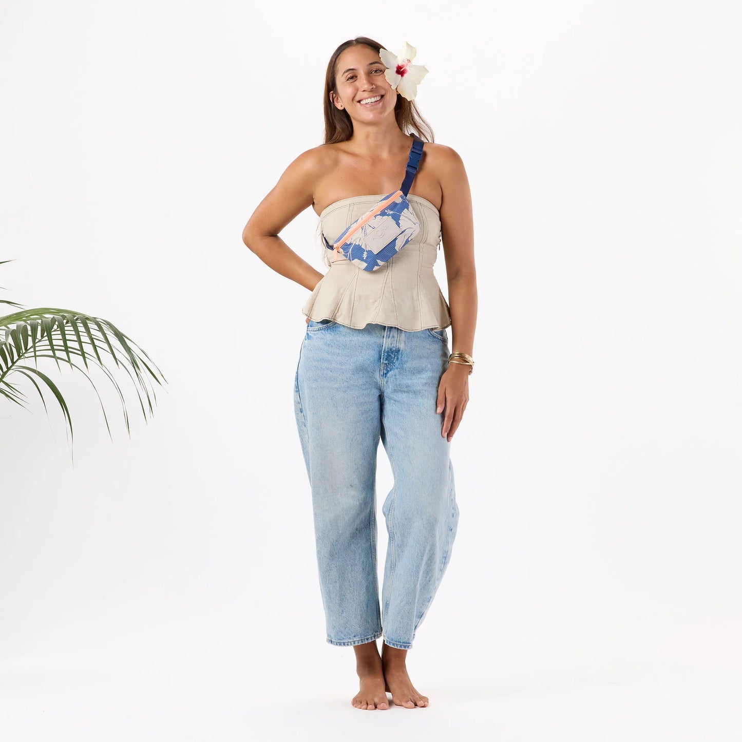 Woman wearing a strapless top and jeans with a floral headpiece and bag, standing against a white background.