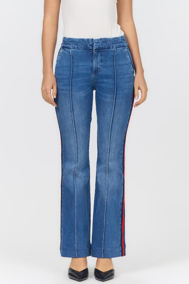 A model is wearing a pair of high-waisted blue denim flare jeans with distinctive red and navy striped side panels, styled with a white top and black heels against a light gray studio background.