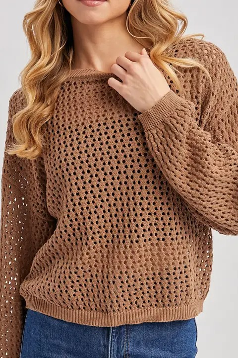 A woman wearing a brown open-knit crewneck sweater with blue jeans, shown from the chin down against a light gray studio background.
