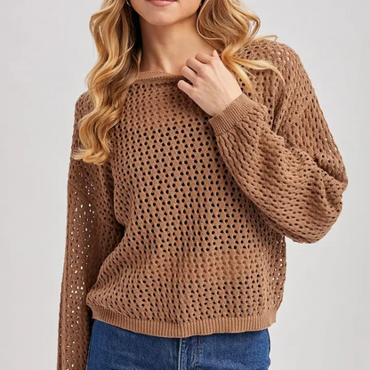 A woman wearing a brown open-knit crewneck sweater with blue jeans, shown from the chin down against a light gray studio background.