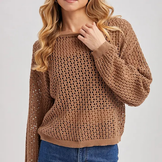 A woman wearing a brown open-knit crewneck sweater with blue jeans, shown from the chin down against a light gray studio background.
