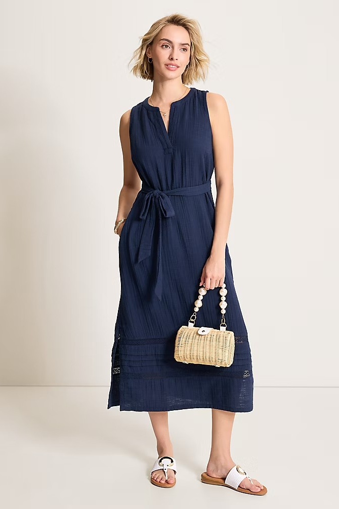 Woman wearing a navy sleeveless dress holding a woven handbag on a plain background