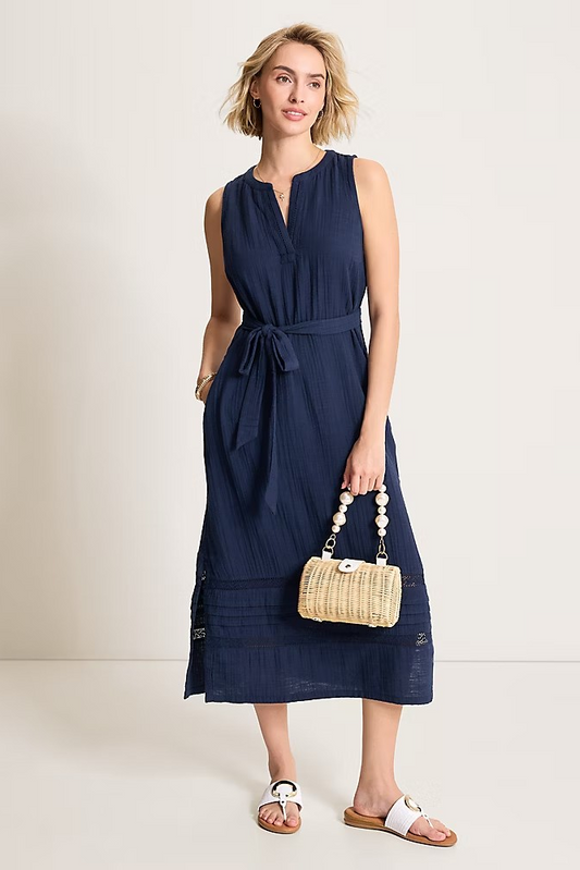 Woman wearing a navy sleeveless dress holding a woven handbag on a plain background