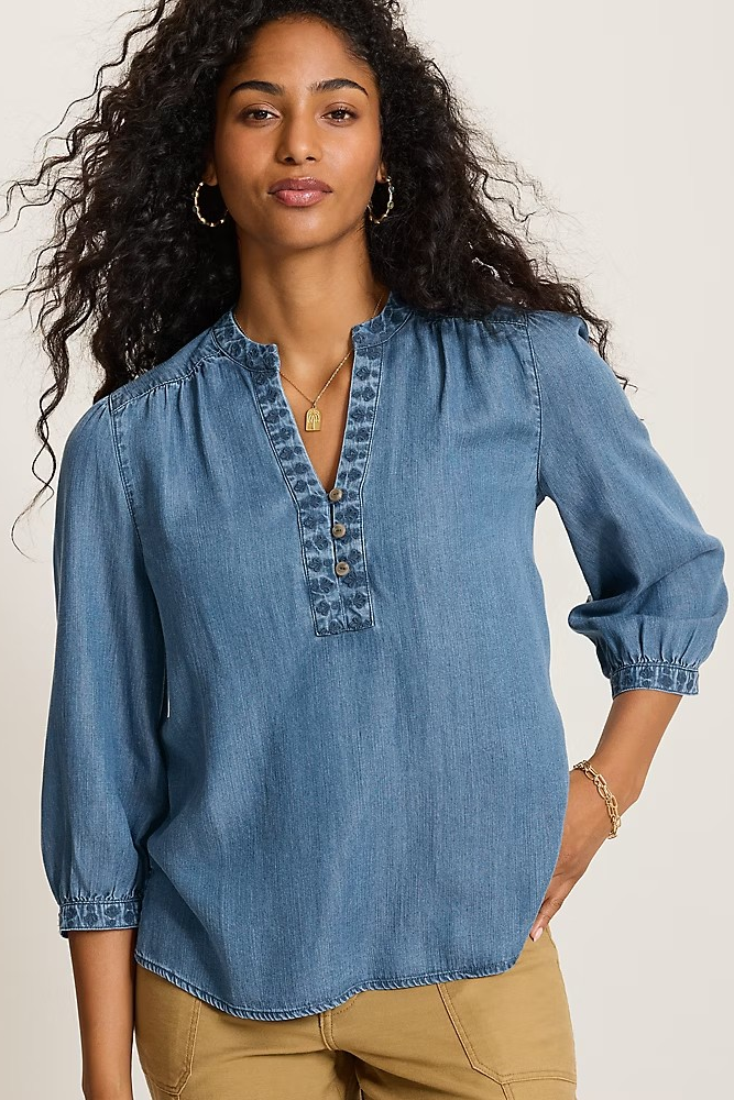 A woman wearing a blue denim long-sleeve blouse with a buttoned split neckline and gathered sleeves, paired with tan pants, against a plain light background.