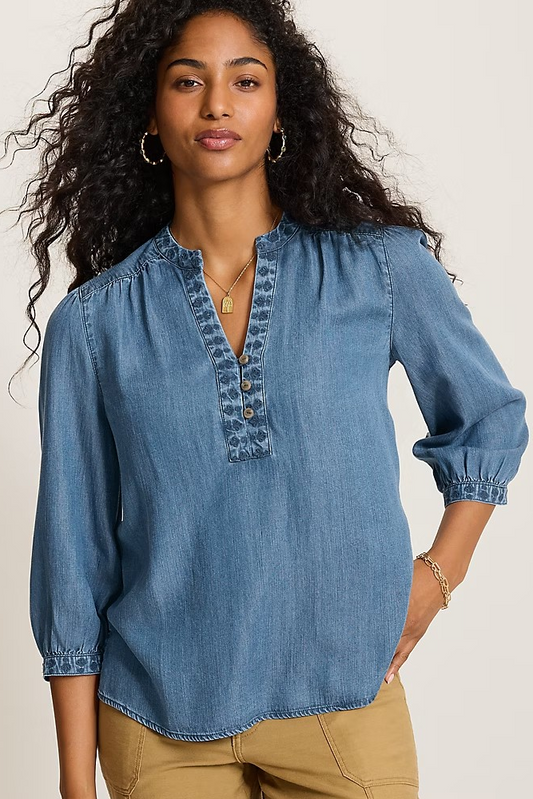 A woman wearing a blue denim long-sleeve blouse with a buttoned split neckline and gathered sleeves, paired with tan pants, against a plain light background.