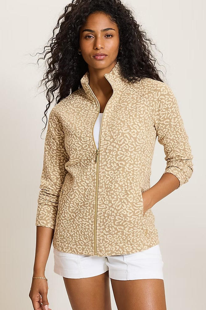 A woman modeling a beige and tan leopard-print zip-up jacket with a stand collar, worn over a white top and paired with white shorts, against a solid light beige background.