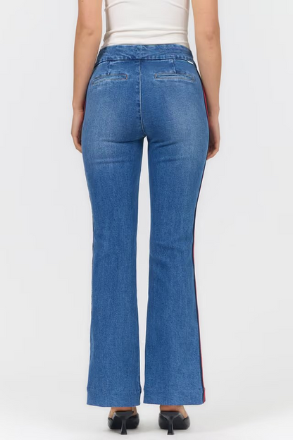 Rear view of a model wearing blue flare jeans with a red and blue side stripe, paired with a white top and black heels, against a light gray studio background.