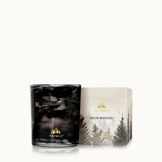 A THYMES Noir Woods scented candle in a dark, smoky black glass jar, shown next to its cream-colored packaging box featuring a gold logo and a pine tree illustration.