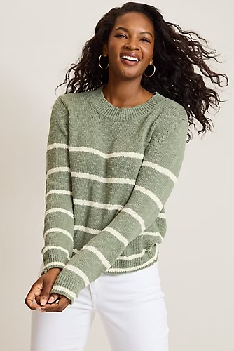 A woman smiling and wearing a sage green knit sweater with horizontal cream stripes, paired with white pants, against a plain light beige studio background.