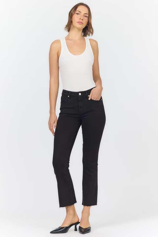 A woman modeling black cropped flare jeans with a white ribbed tank top and black pointed mule heels, standing against a light gray studio background.